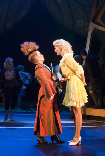 Kaksi teatteriasuihin pukeutunutta naista kohtaa toisensa näyttämöllä; toisella on pitkä, punainen mekko ja dramaattinen tukka, toisella lyhyt, keltainen mekko ja vaaleat kiharat, ja he ovat kohtauksessa, jossa on muita näyttelijöitä ja värikäs lavaste heidän takanaan.