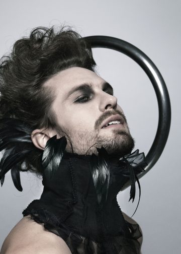 Henkilö, jolla on dramaattinen meikki ja höyhenpeitteiset asusteet, poseeraa harmaata taustaa vasten, ja hänen päänsä takana on musta pyöreä rengas, joka luo halo-vaikutelman.