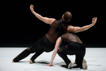 Kaksi nykytanssijaa mustissa, läpikuultavissa puvuissa esiintyy valkoisella näyttämöllä, jossa on tumma tausta. Toinen nojaa taaksepäin kädet ojennettuina, kun taas toinen kyykistyy matalalle ja tukeutuu yhdellä kädellä lattiaan.