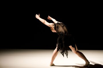 Mustaan mekkoon pukeutunut tanssija esiintyy hämärästi valaistulla näyttämöllä nojautuneena taaksepäin, kädet levällään ja pää kallellaan, luoden dramaattisen ja ilmeikkään poseerauksen tummaa taustaa vasten.