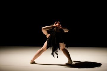 Mustaan pukuun pukeutunut tanssija seisoo valonheittimillä valaistulla näyttämöllä nojautuneena taaksepäin kädet kohotettuna ja sormet yhteenliitettynä, muodostaen dramaattisen, ilmeikkään poseerauksen tummaa taustaa vasten.