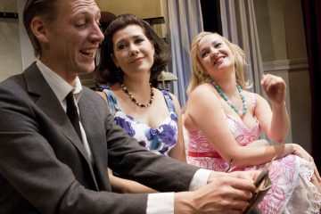 Kolme ihmistä istuu tiiviisti yhdessä sisätiloissa. Vasemmalla istuva pukuun pukeutunut mies hymyilee puhuessaan. Hänen vieressään istuu kaksi värikkäisiin mekkoihin ja kaulakoruihin pukeutunutta naista, jotka molemmat hymyilevät ja katsovat häntä huvittuneesti.