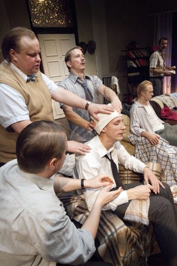 Viisi miestä on kokoontunut viihtyisään huoneeseen, ja osa heistä istuu peittojen peittämillä sängyillä. Kaksi miestä seisoo ja laskee kätensä varovasti istuvan miehen olkapäille ja päähän, mahdollisesti lohduttaen tai hoitaen häntä. Kohtaus näyttää teatraaliselta ja tunteikkaalta.