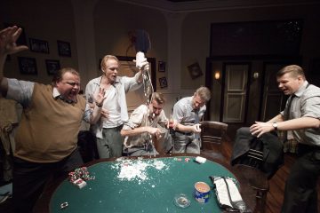 Viisi miestä reagoi dramaattisesti pokeripöydän ympärillä, joka on peitetty roiskuneilla pelimerkeillä ja jauheella, ja yksi mies seisoo ja tarttuu toiseen, mikä luo kaoottisen kohtauksen hämärässä huoneessa.