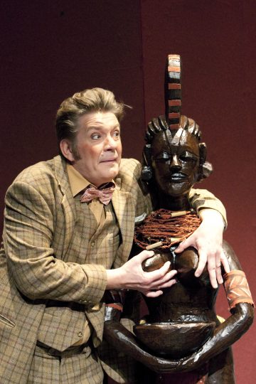 Ruudulliseen pukuun ja rusettisolmioon pukeutunut henkilö poseeraa silmät suurina ja yllättynyt ilme kasvoillaan halaten suurta, tummaa puupatsasta, joka esittää kaulakoruilla ja raidallisella päähineellä koristeltua naista punertavaa taustaa vasten.