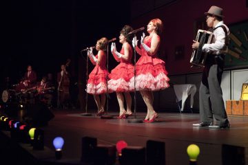 Kolme punavalkoisiin mekkoihin ja hanskoihin pukeutunutta naista laulaa lavalla mikrofoneihin, ja heitä säestävät muusikot, kuten harmonikkaa soittava mies. Lavalla on värikkäitä lamppuja.