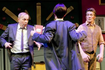 Kolme näyttelijää näyttämöllä esiintyy komediallisessa kohtauksessa; yksi pukumies näyttää yllättyneeltä, toinen ruudullinen mies, jolla on rusetti, näyttää järkyttyneeltä, kun taas kolmas, selkä kameraan päin, pitää kädessään punaista kirjaa, jossa lukee 