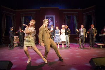 Vilkas lavaesitys, jossa vintage-asuihin pukeutuneet näyttelijät laulavat ja tanssivat; nainen ja mies ovat keskellä ja poseeraavat energisesti, kun muut näyttelijät näyttelevät ja laulavat taustalla.