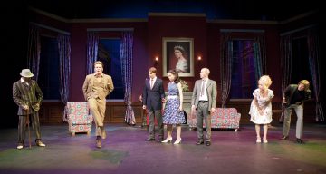 Seitsemän värikkäisiin pukuihin pukeutuneen näyttelijän ryhmä seisoo teatterilavalla, joka on lavastettu olohuoneeksi, jossa on kukkasohvat, kuviolliset verhot ja naisen muotokuva heidän takanaan olevalla seinällä.