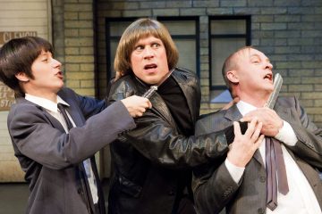 Kolme näyttelijää näyttämöllä dramaattisessa jännityksessä: nahkatakkinen henkilö näyttää hälyttyneeltä, kun yksi pukumies uhkailee häntä aseella ja tarttuu samalla toiseen pukumieheen, joka pitelee puukkoa ensimmäisen henkilön kurkulla.