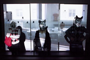 Kolme sudenmaskeihin pukeutunutta ihmistä seisoo kolmen lasipaneelin edessä, joihin on heijastettu luokkahuonekohtaus. Yhdellä henkilöllä on punainen laukku kädessään, ja muut seisovat kädet lanteilla tai sivuillaan.