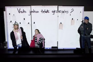 Kolme ihmistä seisoo suuren taulun edessä, johon on teipattu suomenkielistä tekstiä ja papereita. Vasemmalla on kaksi naista, joilla on hattu ja huivi päässään, ja oikealla seisoo siniseen lippikseen pukeutunut mies kädet ristissä.