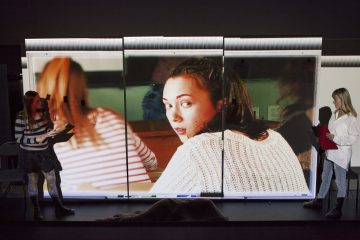 Kaksi ihmistä seisoo suuren, valaistun valkokankaan molemmin puolin, kun lähikuvassa näkyy raitapaitainen nainen, joka kääntyy katsomaan olkansa yli takaisin luokkahuoneessa.