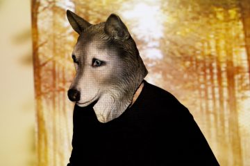 Henkilö, jolla on realistinen susinaamari ja musta paita, seisoo sumean, syksynvärisen metsätaustan edessä.