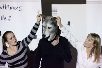 Kaksi naista, joista toisella on raitapaita ja toisella valkoinen paita, ovat vuorovaikutuksessa keskellä olevan henkilön kanssa, jolla on realistinen susinaamari. Toinen nainen pitää naamarin korvaa ylhäällä, kun toinen säätää naamiota. Taustalla näkyy epäselvää kirjoitusta.