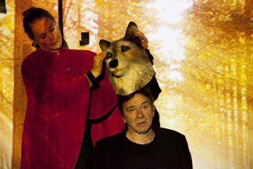 Punaiseen viittaan pukeutunut nainen pitää suurta susinaamiota istuvan miehen pään yläpuolella, taustalla on kultaisia, auringon valaisemia puita, jotka tuovat mieleen metsämaiseman.