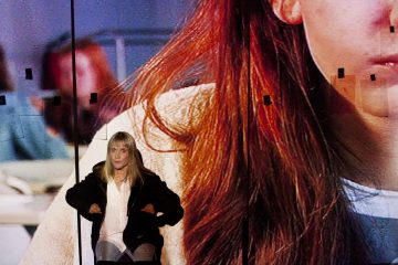 Mustaan takkiin ja valkoiseen paitaan pukeutunut nainen istuu suuren näytön edessä, jossa näkyy kuva pitkät punaiset hiukset omaavasta henkilöstä, jonka kasvot näkyvät osittain. Ruudulle on ripoteltu mustia teipinpaloja.