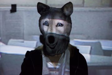 Realistinen susinaamari päällään oleva henkilö seisoo projisoidun taustan edessä, jossa on harmaasävykuvia ja vaakasuoria valoviivoja naamion poikki.