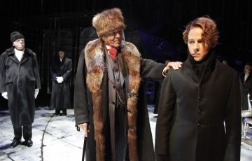 Lavalla seisoo kolme pitkiin, tummiin takkeihin pukeutunutta henkilöä. Keskellä oleva mies, jolla on turkislakki ja turkiskauluksinen takki, lepuuttaa kättään nuoremman, juhlallisen näköisen henkilön olkapäällä. Tausta on hämärästi valaistu.