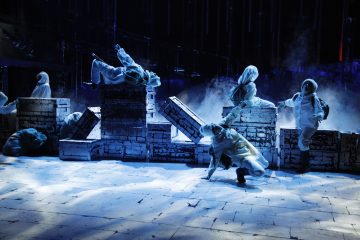 Valkoisiin pukuihin pukeutuneet esiintyjät liikkuvat dramaattisesti hämärästi valaistulla näyttämöllä, jossa on valkoista tiilirakennetta ja hajasumua, mikä luo salaperäisen ja intensiivisen tunnelman sinisessä valaistuksessa.