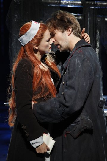 Sairaanhoitajan hattuun pukeutunut nainen ja tummaan takkiin pukeutunut mies seisovat lähekkäin näyttämöllä ja katsovat toisiaan silmiin kiihkeän tunteikkaasti, ikään kuin olisivat aikeissa suudella. Kohtaus on dramaattinen ja teatraalinen.