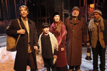 Viisi aikakauden pukuihin pukeutunutta ihmistä seisoo yhdessä lavalla ja katsoo eteenpäin toivon tai yllätyksen ilmein. Taustalla on teatterivalaistus ja lavasteet, jotka viittaavat näytelmän tai musikaalin dramaattiseen kohtaukseen.