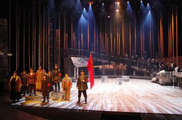 Teatterin näyttämökohtaus, jossa on historiallisiin pukuihin pukeutuneita näyttelijöitä, joista yhdellä on punainen lippu kädessään. Lavastuksessa on puulankkuja, laatikoita ja korotettu kävelytie, ja taustalla on dramaattinen valaistus ja suuri näyttelijäjoukko.