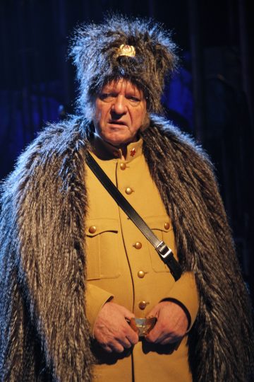 Mies, jolla on turkislakki ja viitta ruskehtavan sotilasasun päällä, seisoo dramaattisessa valaistuksessa ja näyttää vakavalta.