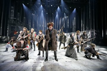 Ryhmä turkisasuihin ja hattuihin pukeutuneita näyttelijöitä esiintyy dramaattisella, hämärästi valaistulla näyttämöllä, heillä on kiväärit kädessään ja he vaikuttavat intensiivisiltä, mikä tuo mieleen historiallisen tai sota-ajan tilanteen.