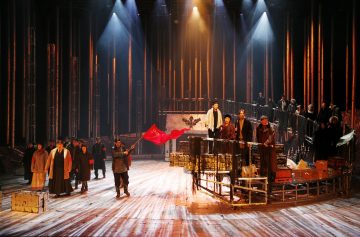 Dramaattinen näyttämökohtaus, jossa on aikakauden pukuihin pukeutuneita esiintyjiä, joilla joillakin on punainen lippu kädessään, ja jonka taustalla on korkeat pystysuorat valot ja puulaatikot, jotka tuovat mieleen historiallisen tai vallankumouksellisen ilmapiirin.