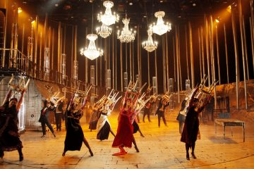 Suuri joukko dramaattisiin pukuihin pukeutuneita esiintyjiä pitää tuoleja korkealla kirkkaasti valaistulla näyttämöllä, jossa on kattokruunuja ja pystysuoria putkia, luoden dynaamisen ja teatraalisen kohtauksen.