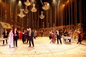 Teatterilava, jossa näyttelijät ovat pukeutuneet tyylikkäisiin aikakauden pukuihin, yläpuolella roikkuvat kattokruunut ja pöydät on katettu juhlallisiin tilaisuuksiin. Kohtaus on vilkas, ja se viittaa tanssiaisiin tai juhliin suurissa puitteissa.