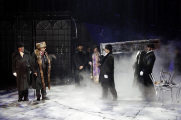 Dramaattinen näyttämökohtaus, jossa tummiin takkeihin ja turkishatuihin pukeutuneet näyttelijät kantavat arkkua sumun läpi. Lavastus on hämärästi valaistu, ja siinä on goottilaisia portteja ja hajallaan olevia tuoleja, jotka luovat synkän, talvisen tunnelman.