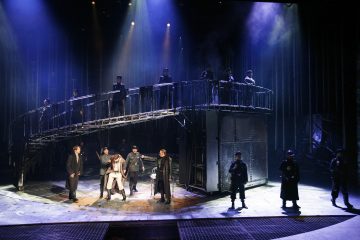 Näyttelijät esiintyvät dramaattisella, hämärästi valaistulla näyttämöllä, jonka yläpuolella on korotettu käytävä ja jonka ylä- ja alapuolella seisoo useita näyttelijöitä, jotka ovat pukeutuneet tummiin, aikakauden tyyliin sopiviin pukuihin tunnelmallisen valaistuksen keskellä.