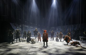 Hämärässä valaistussa dramaattisessa teatterilavalla nähdään joukko näyttelijöitä aikakauden puvuissa. Yksi näyttelijä seisoo näyttämön keskellä punaisena, kun taas muut poseeraavat tai kyyristelevät ympärillä, mikä luo jännittyneen, tunnelmallisen kohtauksen.