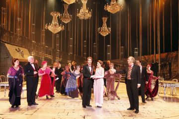 Joukko tyylikkäästi pukeutuneita, aikakauden pukuihin pukeutuneita näyttelijöitä esiintyy suurella näyttämöllä, jossa on kattokruunut, marmorilattia ja kullanvärinen tausta, mikä tuo mieleen teatterituotannon ylenpalttisen tanssisalikohtauksen.