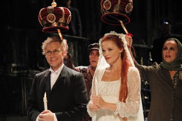 Viralliseen asuun pukeutuneet mies ja nainen seisovat vierekkäin kynttilä kädessään, kun kaksi ihmistä pitää koristeellisia kruunuja heidän päänsä yläpuolella. Naisella on valkoinen mekko, jossa on kukkainen päähine. Tausta on hämärästi valaistu.