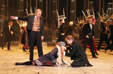 Näyttelijät näyttämöllä esittävät intensiivisen kohtauksen; talvitakkiin ja huiviin pukeutunut nainen polvistuu, kun mustiin pukeutunut mies pitelee häntä, ja muut aikakauden pukuihin pukeutuneet nostavat tuoleja taustalle, mikä luo dramaattisen teatterihetken.