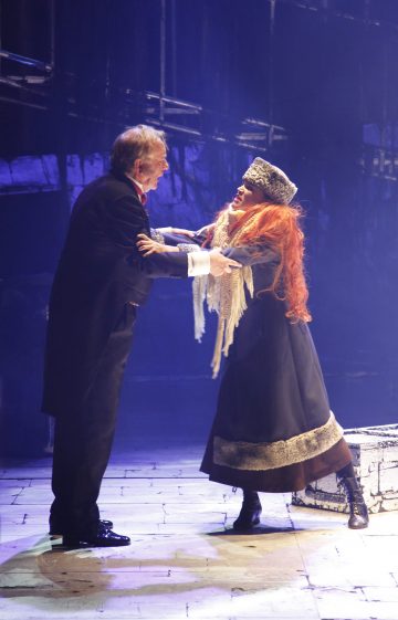 Mustaan pukuun pukeutunut vanhempi mies pitää kädestä naista, jolla on pitkä musta mekko, turkislakki ja oranssi peruukki lavalla, ja molemmilla on tunnepitoinen hetki dramaattisessa valaistuksessa.