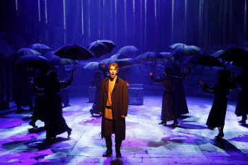 Mies seisoo näyttämön keskellä ja laulaa dramaattisessa valaistuksessa, kun tummiin vaatteisiin pukeutuneet hahmot, joilla on mustat sateenvarjot, ympäröivät häntä teatteriesityksessä. Violetit ja siniset valot luovat tunnelmallista tunnelmaa.