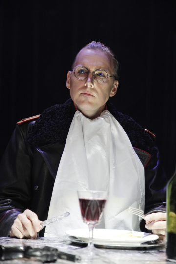 Silmälasipäinen henkilö, jolla on musta turkistakki ja turkiskaulus, istuu pöydän ääressä, valkoinen lautasliina kauluksessa, veitsi ja haarukka kädessä. Pöydällä on lasi punaviiniä ja vihreä pullo.