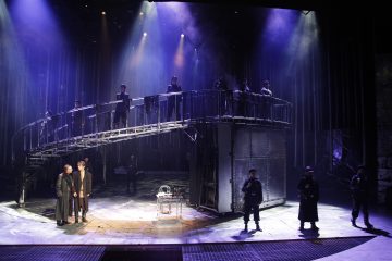 Dramaattinen näyttämökohtaus, jossa tummiin pukuihin pukeutuneet näyttelijät seisovat kaarevalla metallilavalla ja toiset näyttämöllä alla, ja jossa on tunnelmallinen violetti ja sininen valaistus, joka viittaa intensiiviseen teatteriesitykseen.