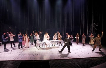 Lavalla esiintyy suuri joukko näyttelijöitä, joista monet ovat pukeutuneet perinteisiin tai vanhoihin pukuihin. Keskeiset hahmot istuvat pitkän pöydän ääressä, ja muut seisovat tai tanssivat heidän ympärillään dramaattisen valaistuksen alla. Kohtaus viittaa vilkkaaseen juhlaan tai kokoontumiseen.