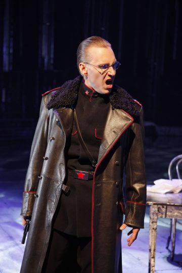 Lavalla seisoo henkilö, jolla on musta sotilastyylinen takki, jossa on punainen reunus ja silmälasit, ja hän näyttää huutavan. Hänellä on ase kädessään ja hän seisoo metallipöydän vieressä, jossa on papereita, dramaattisessa valaistuksessa.