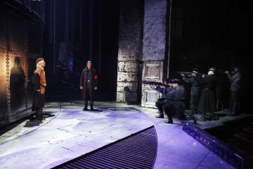 Dramaattisessa näyttämökohtauksessa yksi mies seisoo metalliseinää vasten, kun toinen sotilaspukuun pukeutunut mies kohtaa hänet. Oikealla puolella joukko sotilaita tähtää kiväärillä seinää vasten olevaa miestä. Valaistus on tumma ja voimakas.