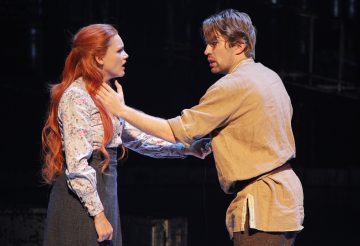 Nainen, jolla on pitkät punaiset hiukset ja kukkapusero, seisoo vastapäätä miestä, jolla on ruskehtava paita. Mies pitää naista olkapäistä kiinni, ja molemmat näyttävät tunteikkailta kuin näyttämöllä tapahtuvassa dramaattisessa kohtauksessa. Tausta on tumma ja epämääräinen.