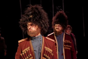 Lavalla seisoo kaksi miestä, joilla on koristeelliset punaiset univormut ja suuret mustat turkishatut, ja he näyttävät vakavilta. Tausta on tumma, ja heidän takanaan näkyy osittain toinen henkilö.