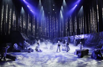 Dramaattinen näyttämökohtaus, jossa harmaisiin pukuihin pukeutuneet näyttelijät liikkuvat hajallaan olevien laatikoiden ja lavasteiden keskellä, ja jota valaisee sininen ja violetti valaistus, jonka taustalla on pystysuoria valonsäteitä, jotka luovat salaperäisen tunnelman.