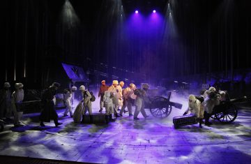 Aikakauden pukuihin pukeutuneet esiintyjät työntävät kärryjä ja kantavat koreja hämärästi valaistulla näyttämöllä, jossa on dramaattinen violetti valaistus ja sumua, mikä viittaa historialliseen tai teolliseen kohtaukseen teatterituotannossa.