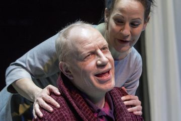 Hymyilevä mies ruskeankeltaisessa kaapussa istuu, kun harmaaseen pukeutunut nainen seisoo hänen takanaan kädet miehen olkapäillä, ja molemmat vaikuttavat iloisilta ja keskustelevilta.